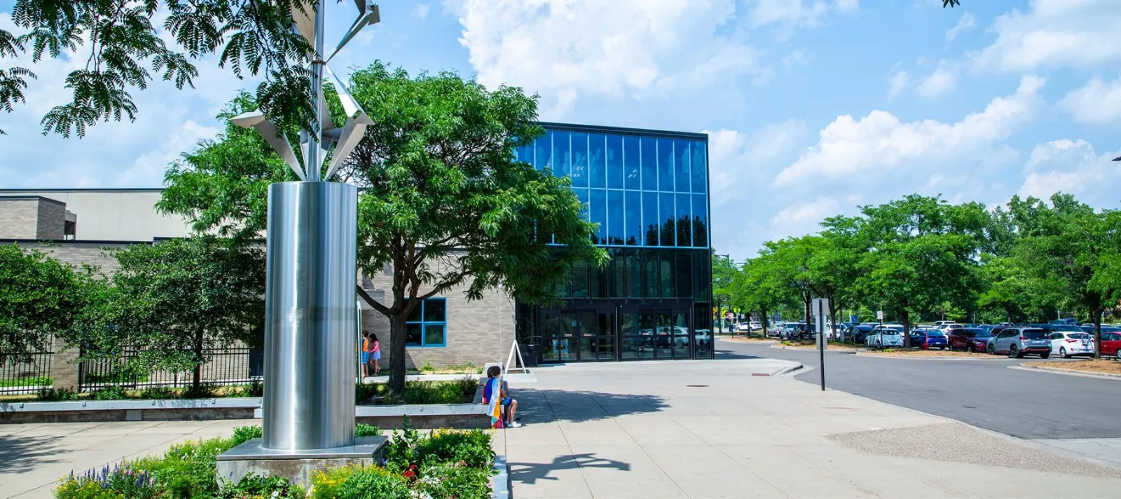 El bloque de entrada de vidrio y los muros de piedra del Rec Center detrás de una escultura de acero y jardineras, con el estacionamiento más allá. — St. Louis Park Rec Center