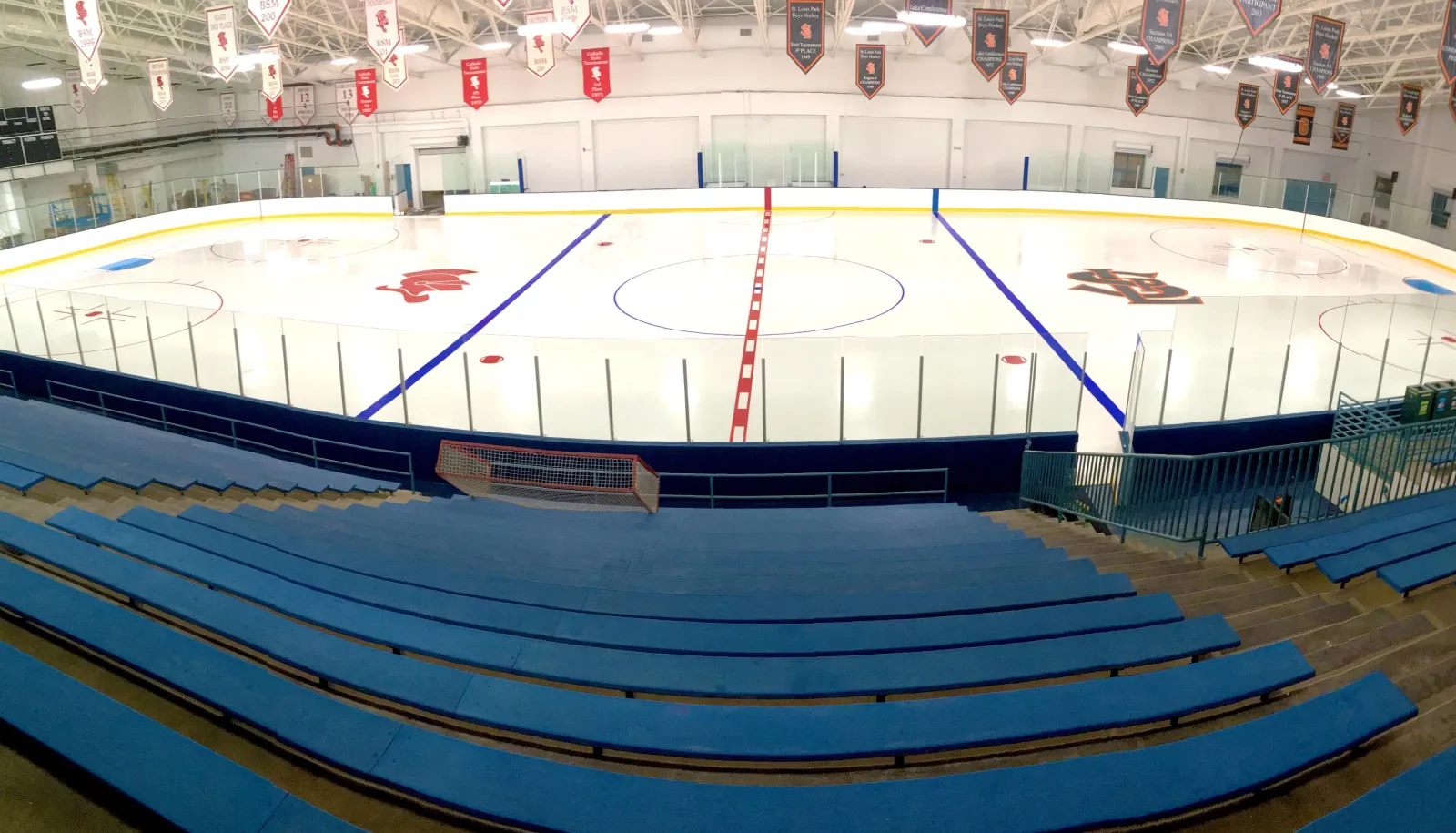 La arena oeste vacía desde lo alto de las gradas azules, con el logotipo del centro de la pista e hileras de mantas colgadas del techo. — St. Louis Park Rec Center