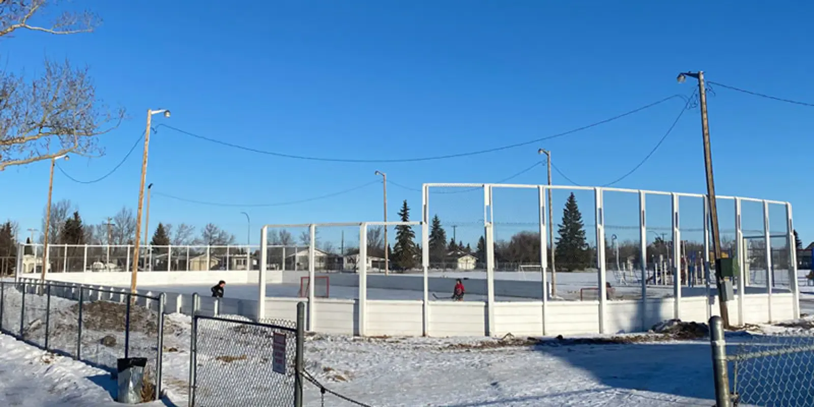 La pista exterior St. Pat's en Camrose bajo un cielo azul de invierno, con una superficie de hielo con vallas y redes alrededor y una hilera de árboles sin hojas detrás. — St. Pat's Rink