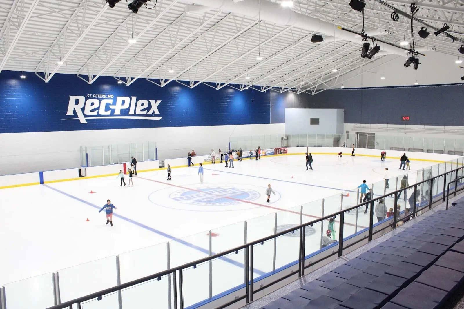 Una sesión de patinaje público en la pista del Rec-Plex, con patinadores repartidos por el hielo detrás de un barandal negro bajo un luminoso techo arqueado. — St. Peters Rec-Plex
