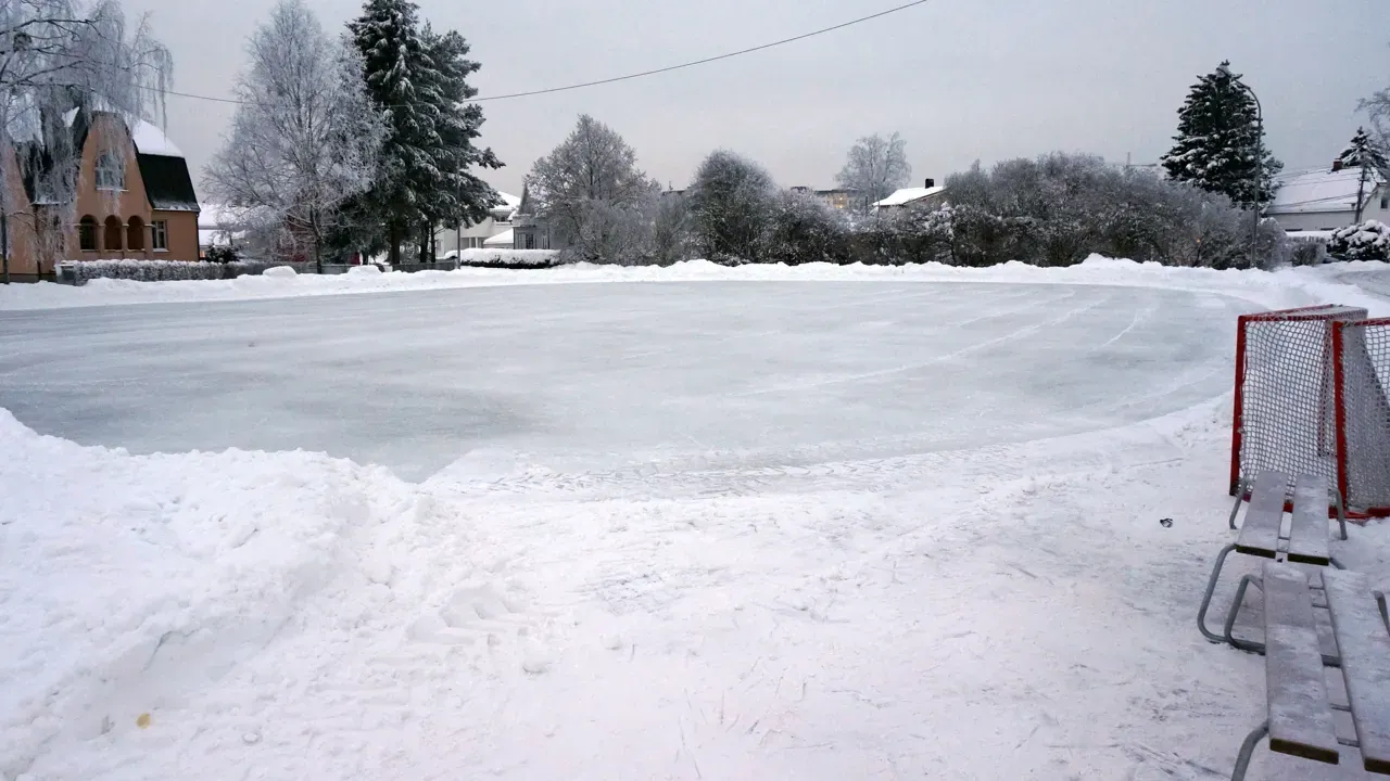 The ice at St. Sunnivas plass in winter, with a hockey goal and a bench at the edge, snow banks and a brick building behind. — St. Sunnivas plass skøytebane (Lillestrøm)