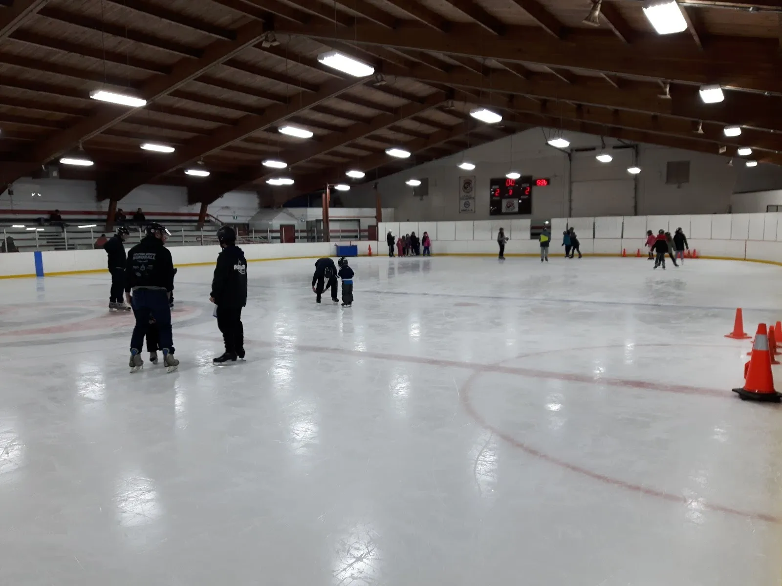 Una sesión de patinaje público en una pista amplia, con unos cuantos patinadores de pie entre conos naranjas bajo un techo con vigas de madera y un marcador en la pared del fondo. — St. Vital Centennial Arena
