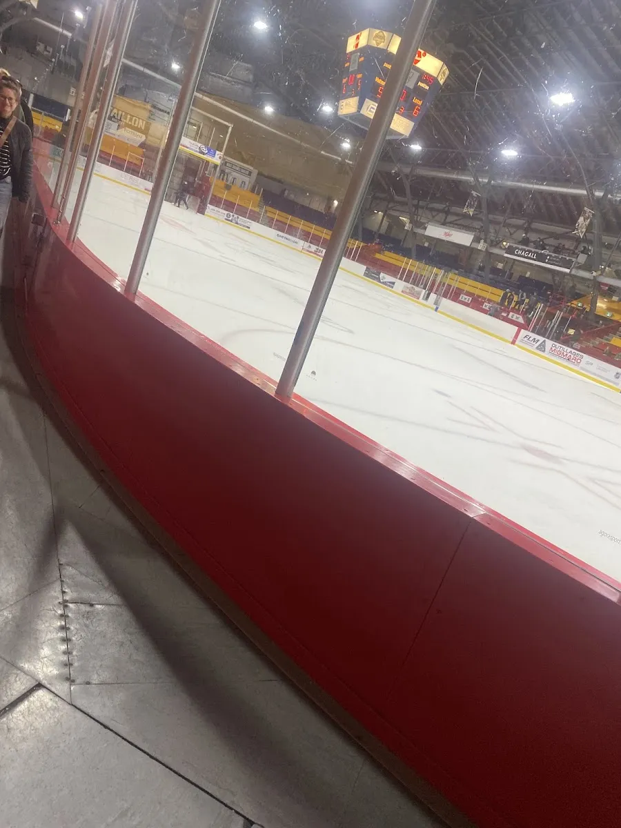 La glace vide vue derrière les bandes rouges et la baie vitrée, avec le tableau indicateur suspendu au toit sombre et les gradins au fond. — Stade L.-P.-Gaucher