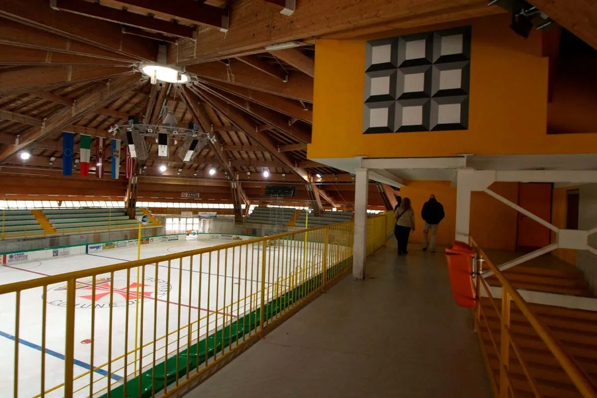 The empty ice of the Cavalese ice stadium with the municipal crest at centre ice, seen from the boards — Stadio del Ghiaccio Cavalese