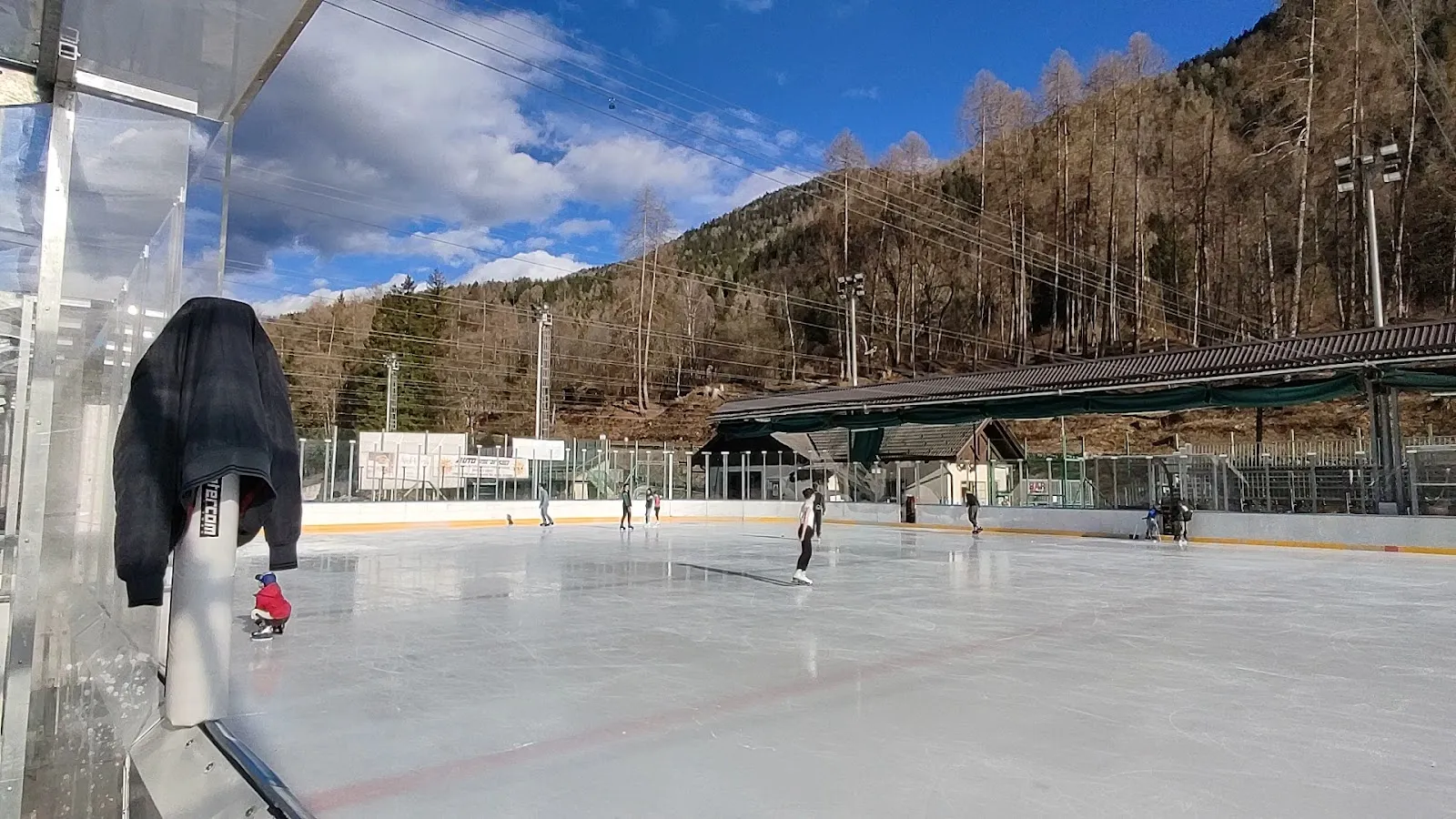 The open-air ice rink at Malé with skaters in the distance and the Val di Sole slopes beyond — Stadio del ghiaccio di Malé