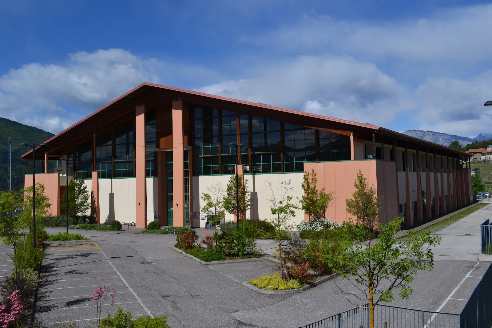 Il lungo edificio in legno e vetro dello stadio del ghiaccio, con il tetto a falde basse, visto dal parcheggio con le montagne sullo sfondo. – Stadio del Ghiaccio Pergine Valsugana