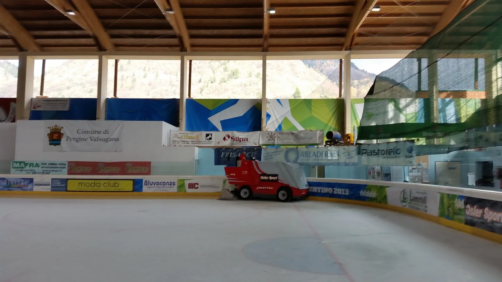 Il ghiaccio vuoto al coperto con una sgombraneve rossa ferma a bordo pista, la vetrata laterale e il pannello del Comune di Pergine Valsugana sulla balaustra. – Stadio del Ghiaccio Pergine Valsugana