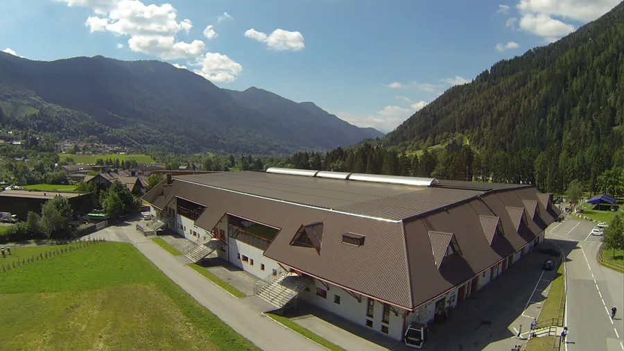 An aerial view of the long brown-roofed Pinzolo ice stadium with its dormer windows, set on a green meadow with wooded mountains behind and a road passing alongside. – Pinzolo Ice (Stadio del ghiaccio)