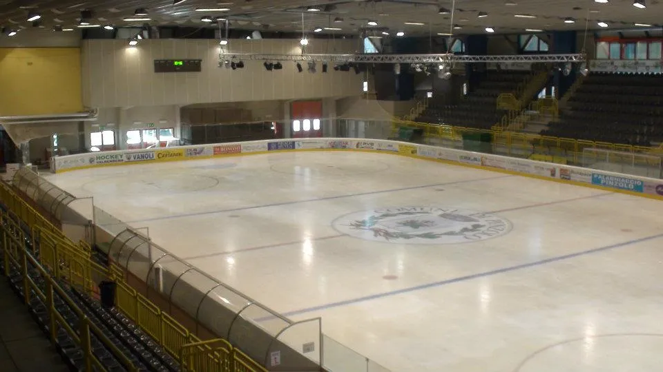 The empty ice sheet of the Pinzolo stadium seen from the stands, with the centre-circle emblem, yellow railings, advertising boards and the covered stands on the far side. – Pinzolo Ice (Stadio del ghiaccio)