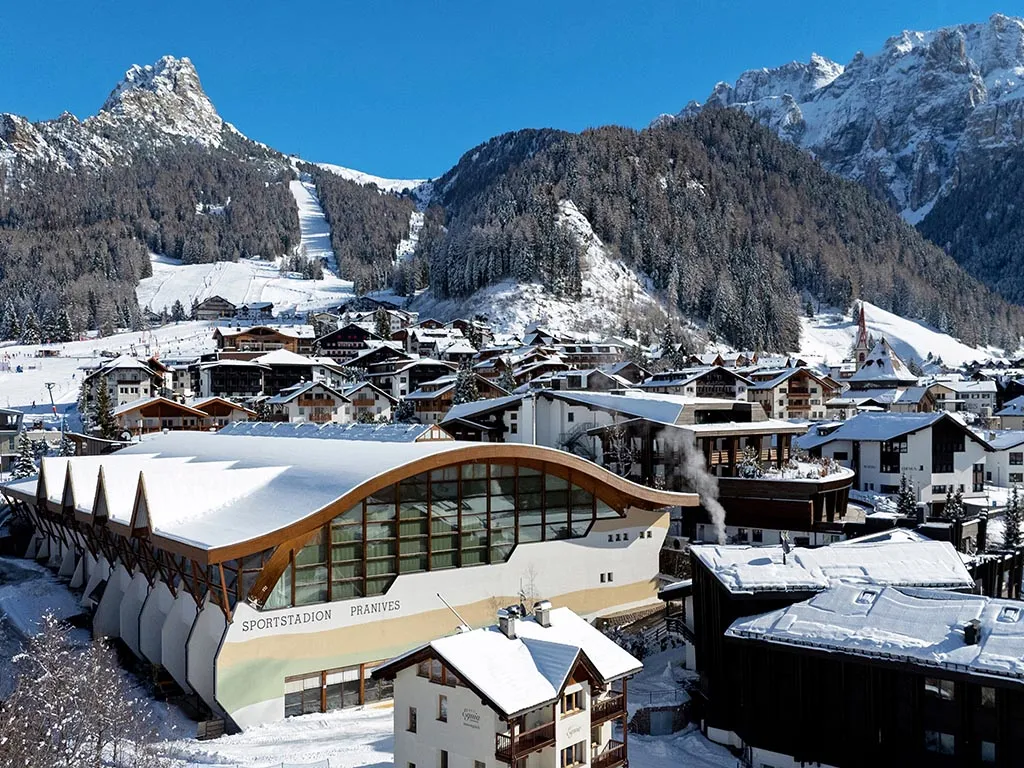 The curved-roof Pranives ice stadium among the snow-covered houses of Selva with the Dolomite peaks behind. — Stadio del ghiaccio Pranives