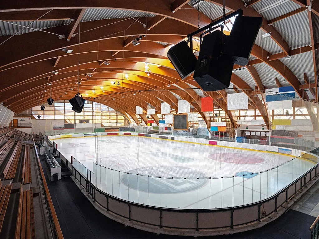 The empty white ice sheet of the Pranives stadium under its curved laminated-timber roof, with the stands along the boards. — Stadio del ghiaccio Pranives