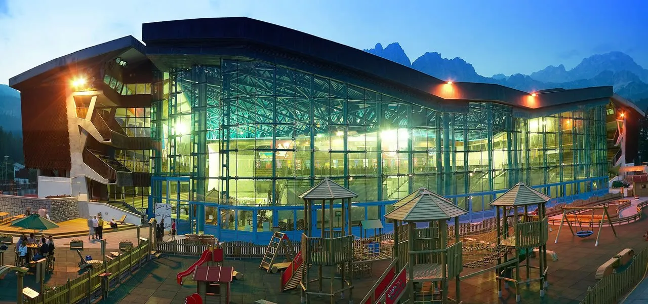 The glass-fronted Olympic Ice Stadium of Cortina lit at dusk with the Dolomite peaks behind and a children's playground in the foreground. — Stadio Olimpico del Ghiaccio (Cortina d'Ampezzo)