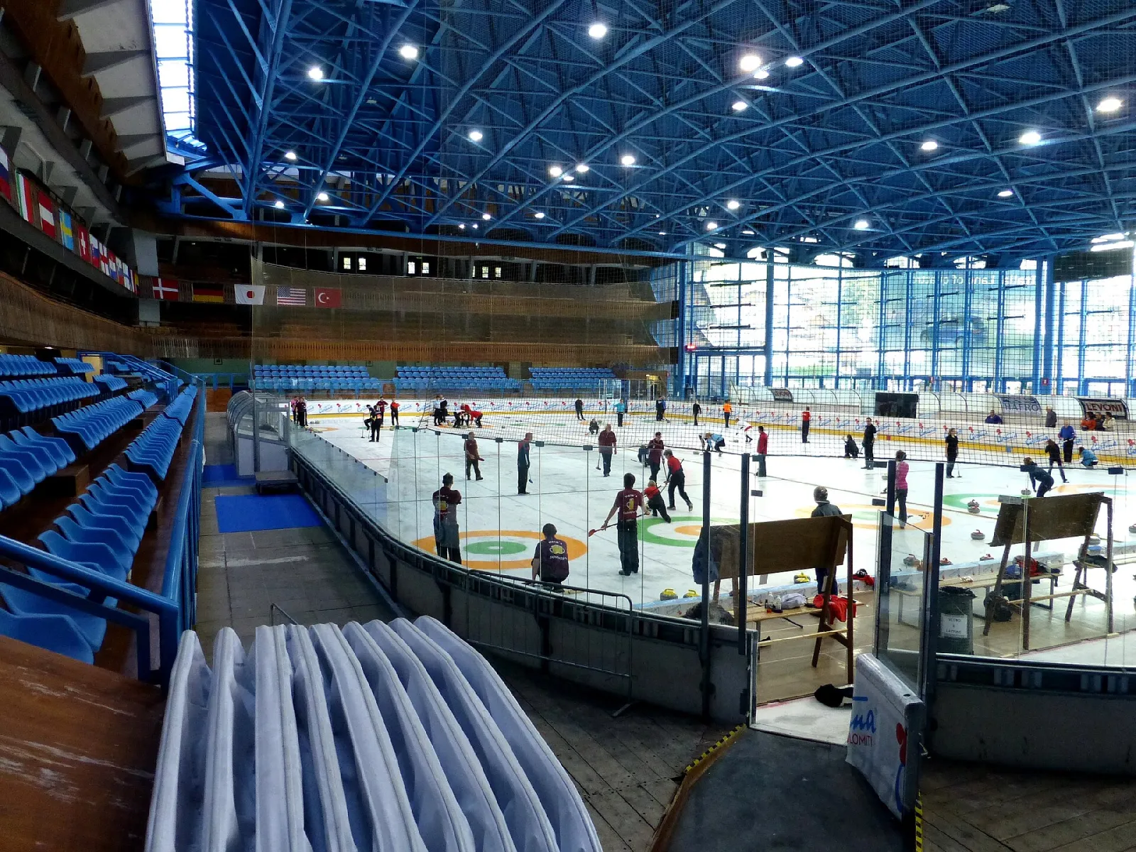 Inside the stadium under its blue steel roof, with a curling event on the ice, the stands on the left and the glass wall on the right. — Stadio Olimpico del Ghiaccio (Cortina d'Ampezzo)