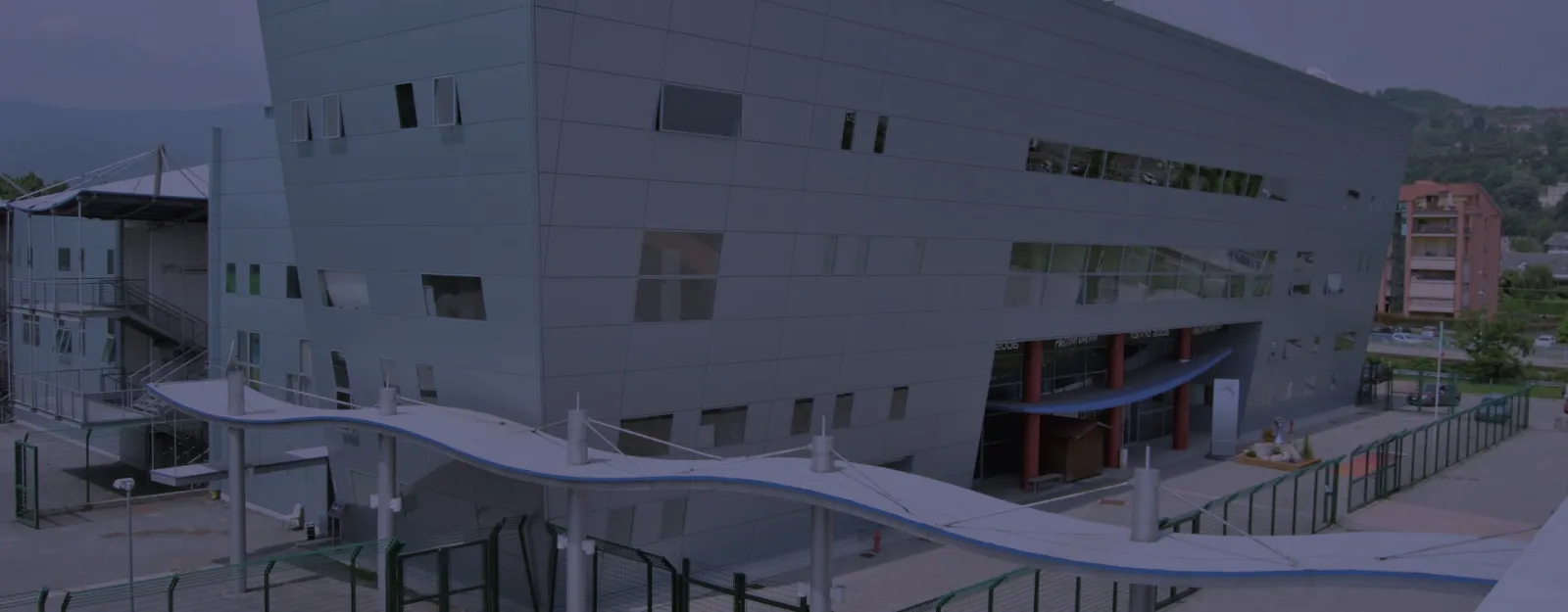 La facciata grigia e spigolosa dello Stadio Olimpico del Ghiaccio con la tettoia curva sopra l'ingresso, con la scritta Torino 2006, vista dal piazzale. – Stadio Olimpico del Ghiaccio (Ice Pole Pinerolo)
