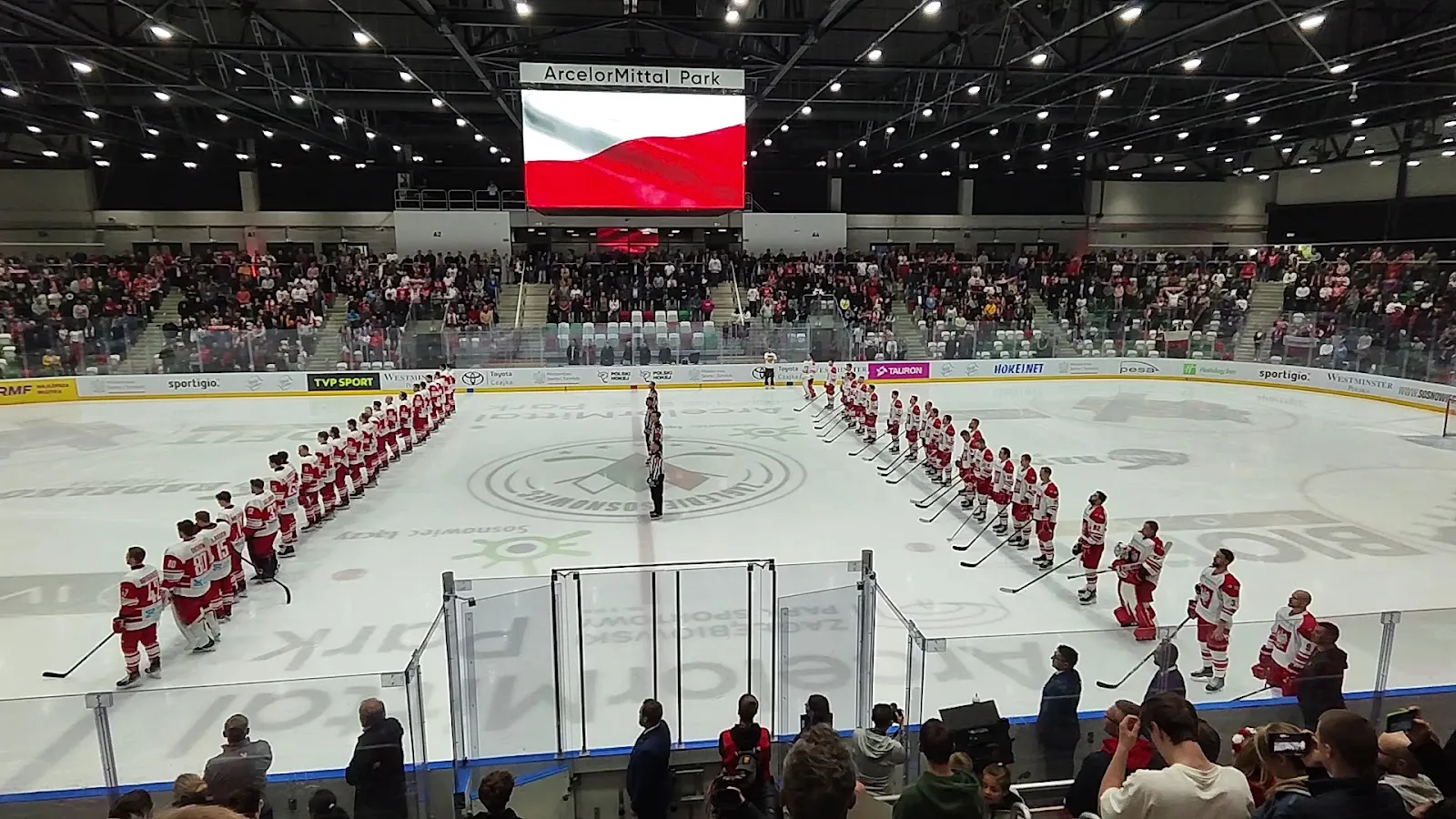 Ice hockey teams lined up on the ice at the Sosnowiec winter stadium before a match, with the spectator stands behind — Stadion Zimowy Sosnowiec (Zagłębiowski Park Sportowy)