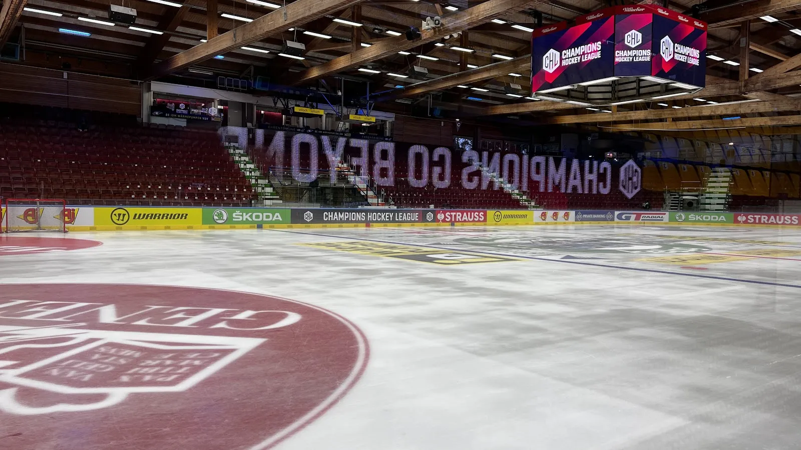 Das leere Eis der Stadthalle Villach auf Eisniveau, mit dem roten Wappen in der Eismitte, den Banden der Champions Hockey League und den leeren Rängen dahinter – Stadthalle Villach
