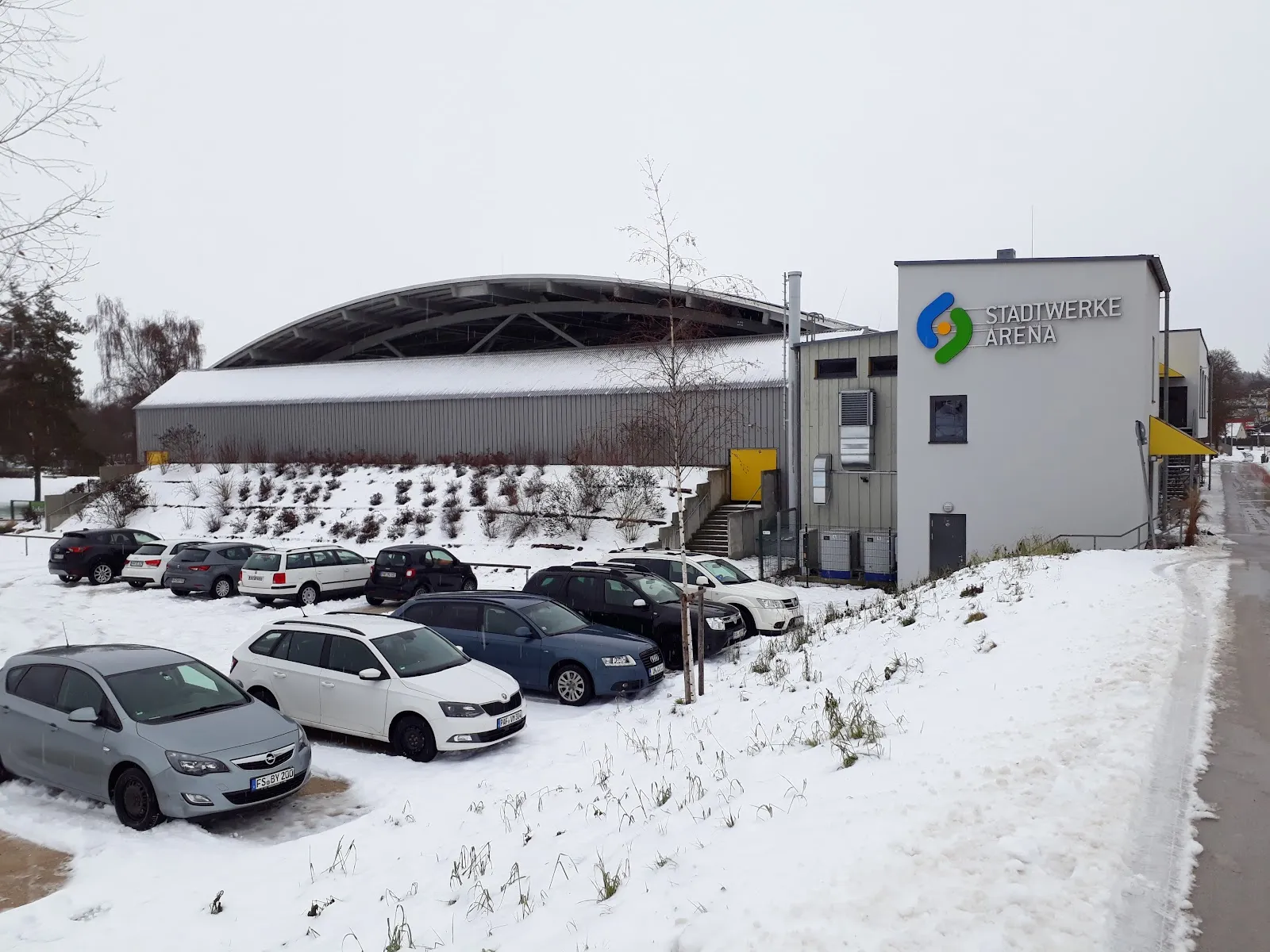 The arched roof of the hall beside the grey block carrying the Stadtwerke Arena sign, with a car park in front under a dusting of snow. — Stadtwerke-Arena Pfaffenhofen