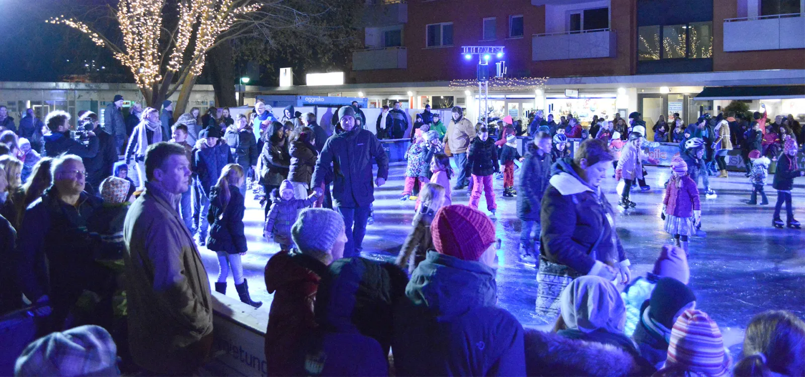 Schlittschuhläufer und Zuschauer an den Banden der Elmshorner Eisbahn bei Nacht, mit den Gebäuden am Bahnhof dahinter und einem Baum mit Lichterketten. – Stadtwerke Eisvergnügen Elmshorn