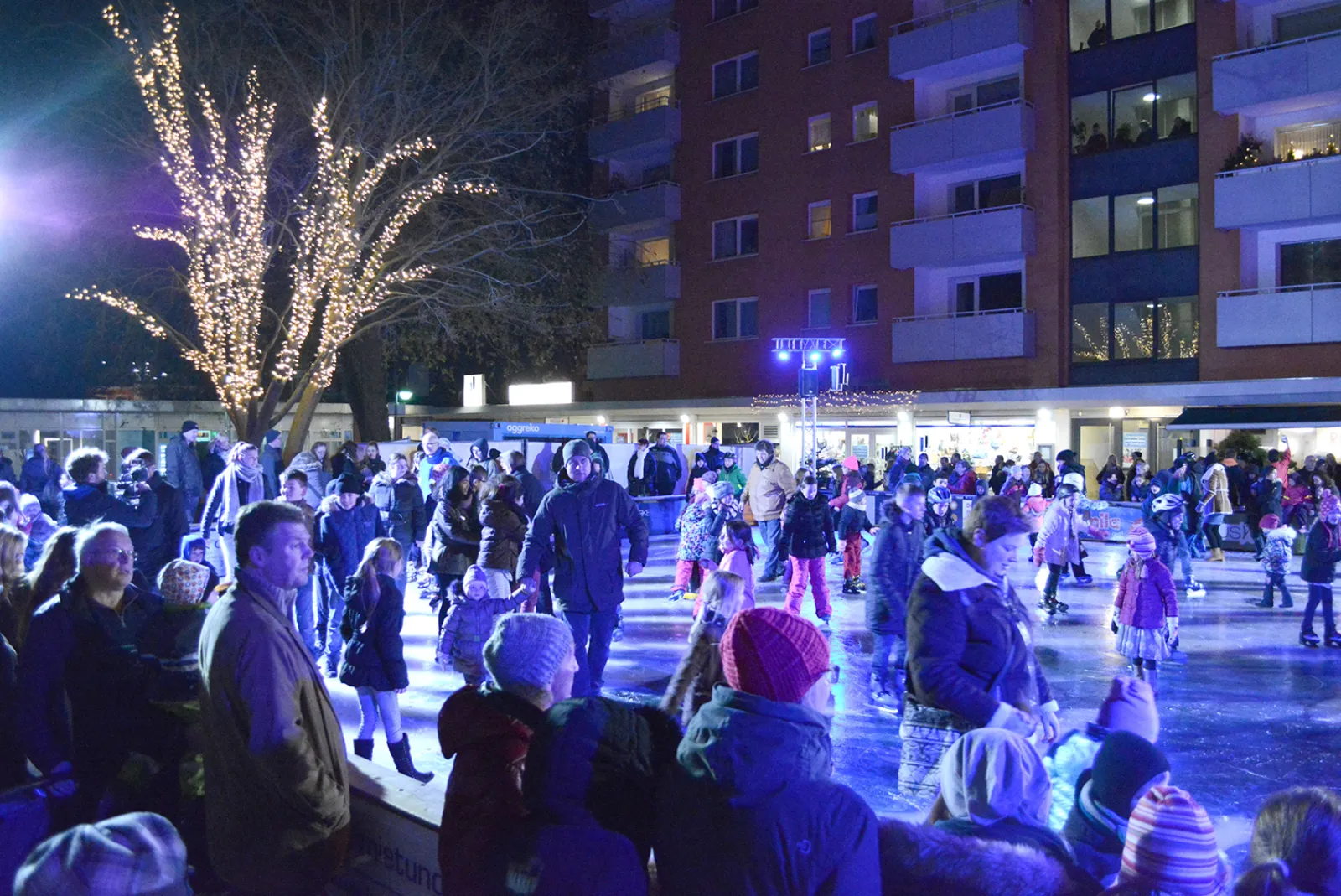 Die Freiluft-Eisbahn am Bahnhof Elmshorn am Abend, voller Schlittschuhläufer unter blau-weißer Beleuchtung, mit einem Wohnblock dahinter. – Stadtwerke Eisvergnügen Elmshorn