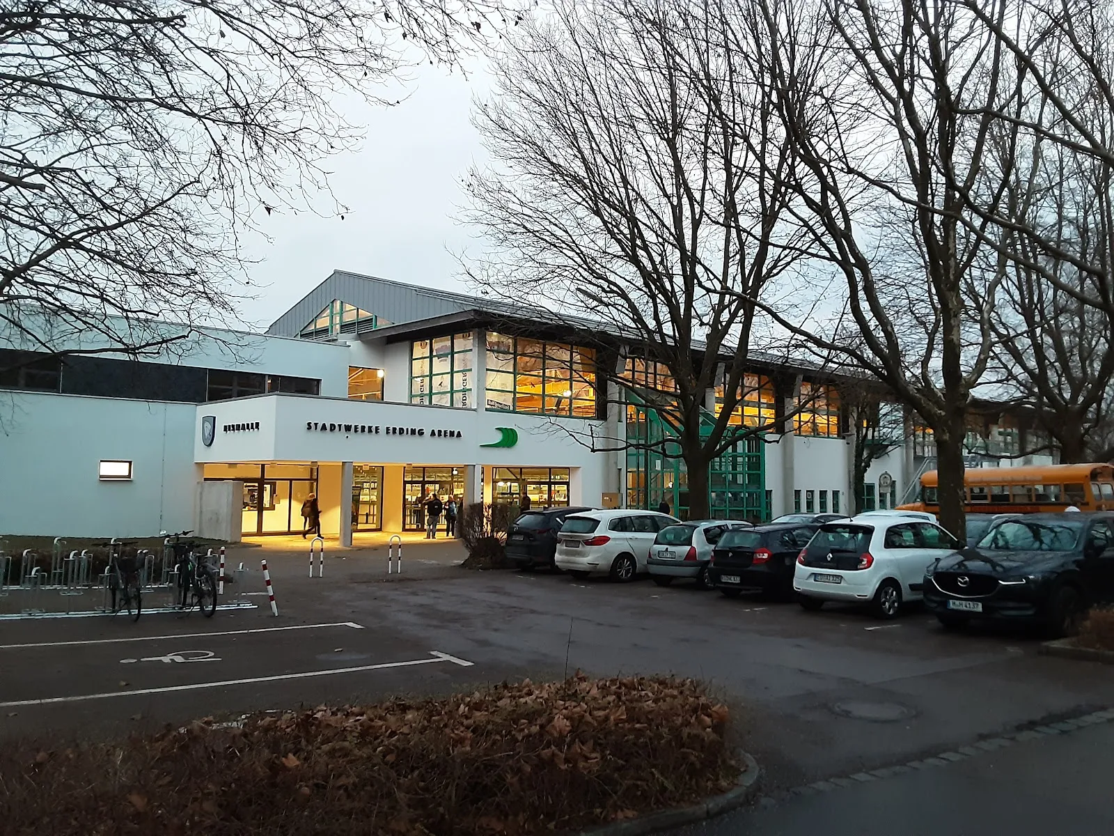 Der beleuchtete Eingang der Arena in der Dämmerung, ein weißes Gebäude mit großen Glasfronten und dem Schriftzug Stadtwerke Erding Arena, davor parkende Autos und kahle Bäume ringsum. – Stadtwerke Erding Arena - Eishalle