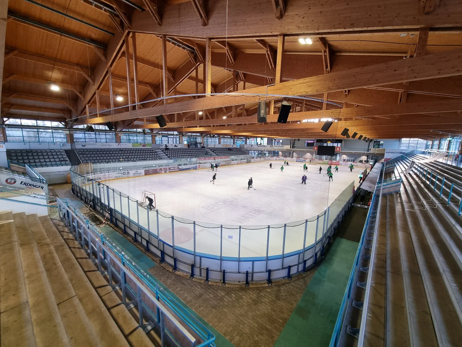 Die Eisfläche, von den steilen Rängen unter der Holzleimbinder-Dachkonstruktion der Halle aus gesehen, mit einigen Spielern beim Training auf der gegenüberliegenden Seite. – Stadtwerke Erding Arena - Eishalle