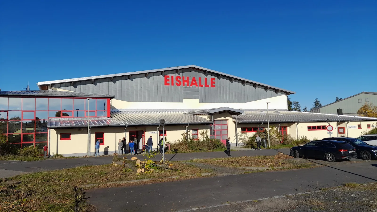 Die niedrige Front der Halle mit dem roten Schriftzug EISHALLE über einem verglasten Eingang, gesehen über den Parkplatz unter tiefblauem Himmel. – Städtische Eishalle Mitterteich