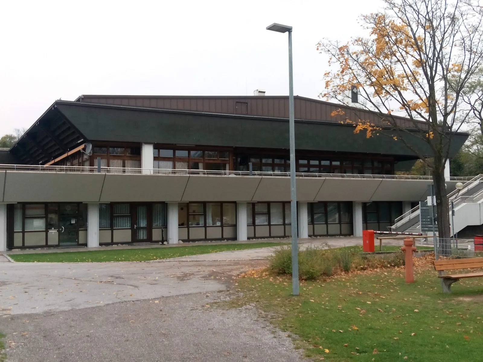The long low Eissporthalle in Landsberg am Lech with its dark sloping roof over the glazed ground floor, seen across the lawn — Städtische Eissporthalle Landsberg