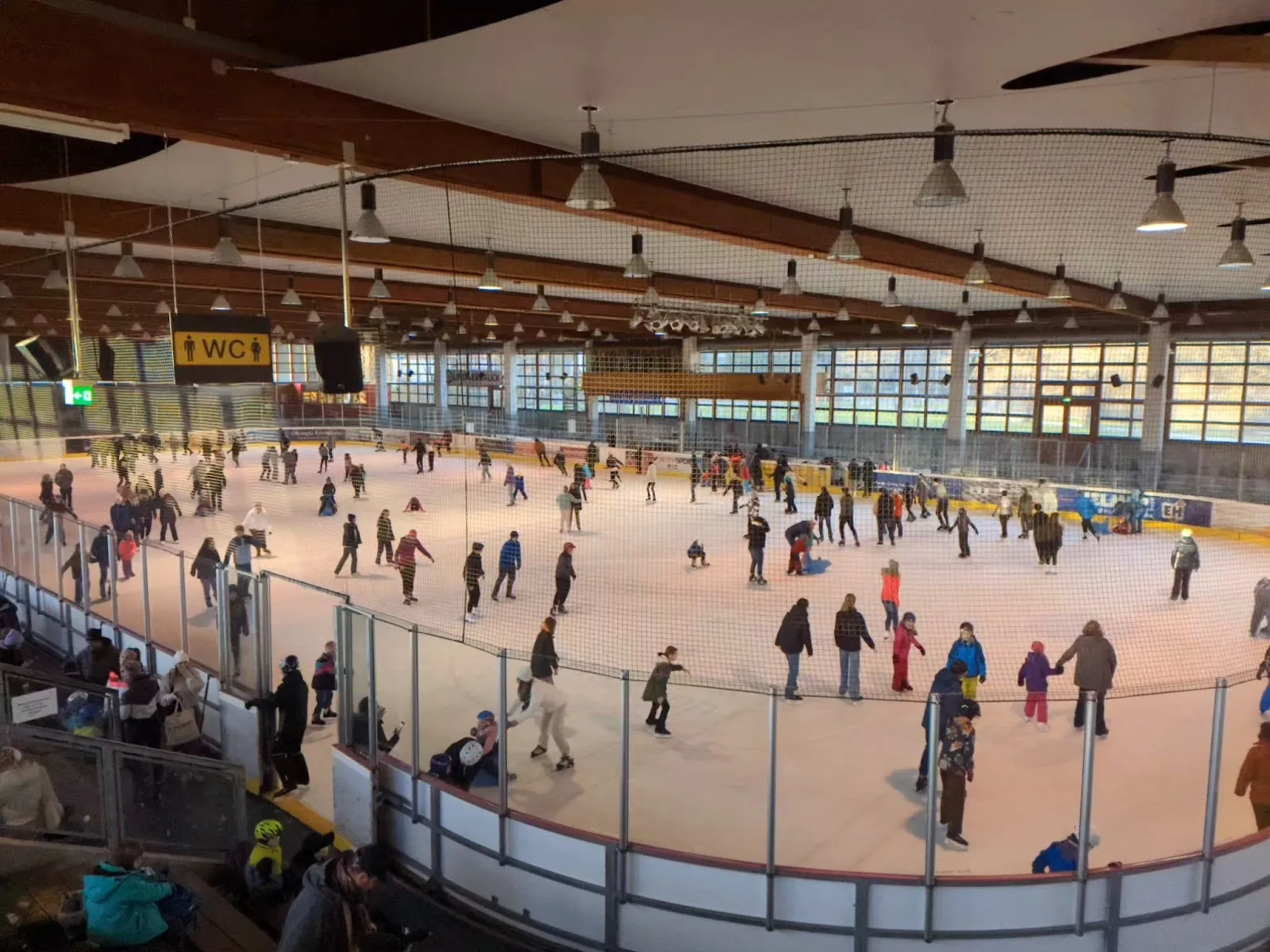 A busy public skating session on the ice of the Landsberg ice hall, with skaters across the whole sheet, the timber roof and the glazed upper wall behind. — Städtische Eissporthalle Landsberg