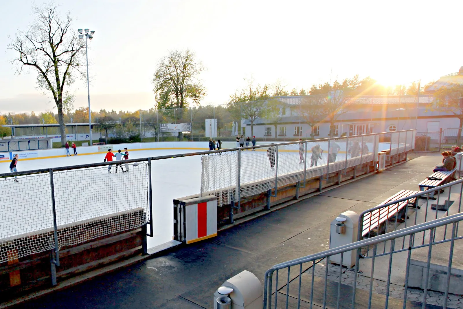 Die Freiluftbahn in der tiefstehenden Abendsonne, die eingezäunte Eisfläche mit einigen Läufern und dahinter die kahlen Bäume und Flutlichtmasten des Parks. – Städtische Kunsteisbahn Dachau
