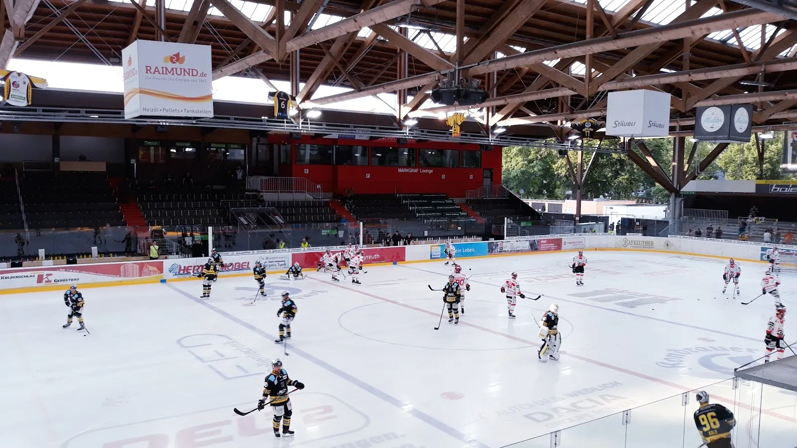 Schlittschuhläufer auf dem Eis des Bayreuther Kunsteisstadions, über die Fläche gesehen, unter dem Dach aus Holz und Stahl mit den Sponsorenbanden dahinter. – Städtisches Eisstadion Bayreuth