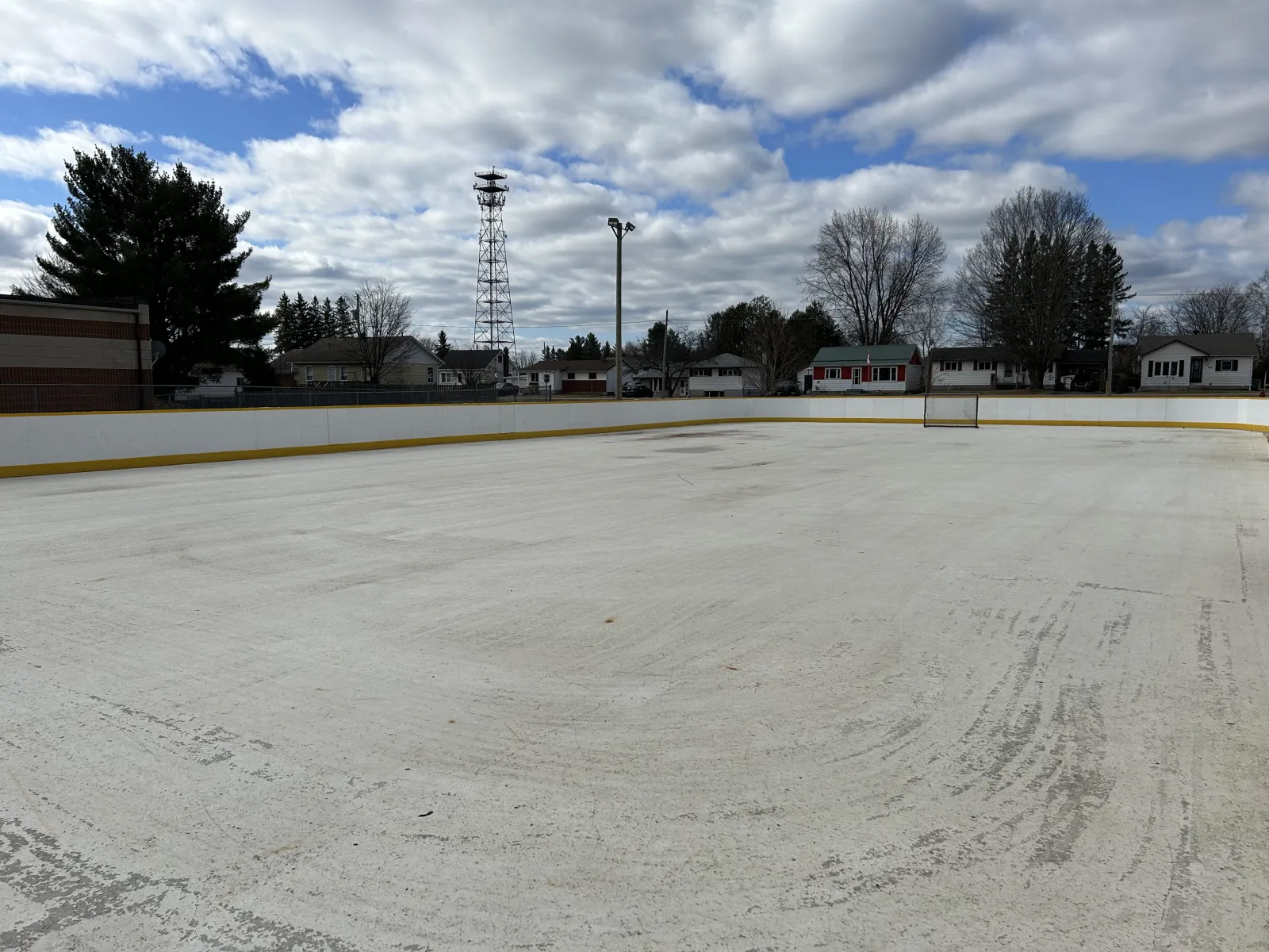 The empty boarded outdoor rink at Stafford Park under a cloudy sky, with a floodlight pole, a radio mast and houses across the street. — Stafford Park Outdoor Rink (Laurentian Valley)