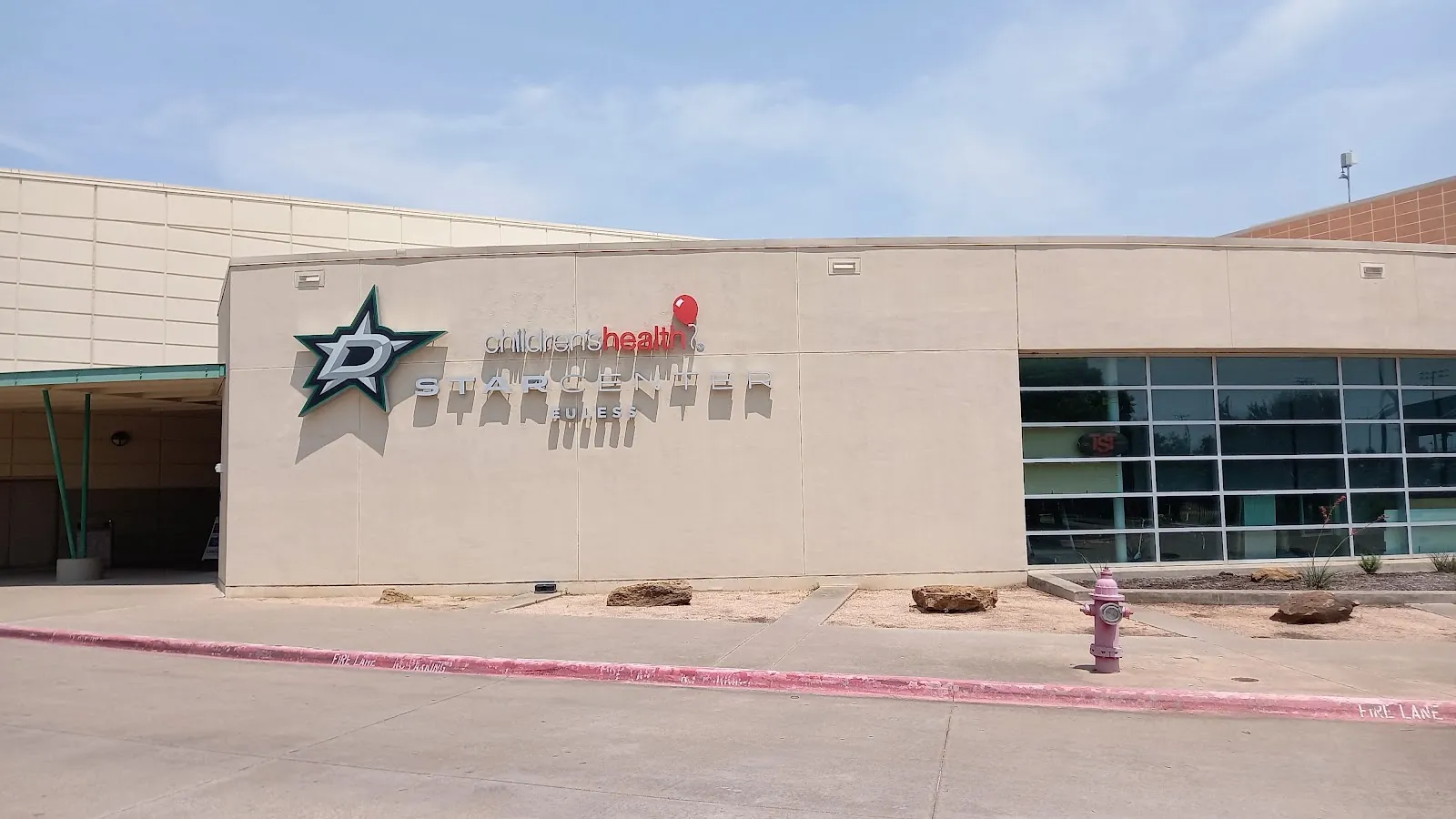 Front of the building: tan facade with the Dallas Stars 'D-star' logo and 'children's health STARCENTER EULESS' wall lettering above the glass entrance, pink fire hydrant on the curb, clear daytime sky. — Children's Health StarCenter Euless