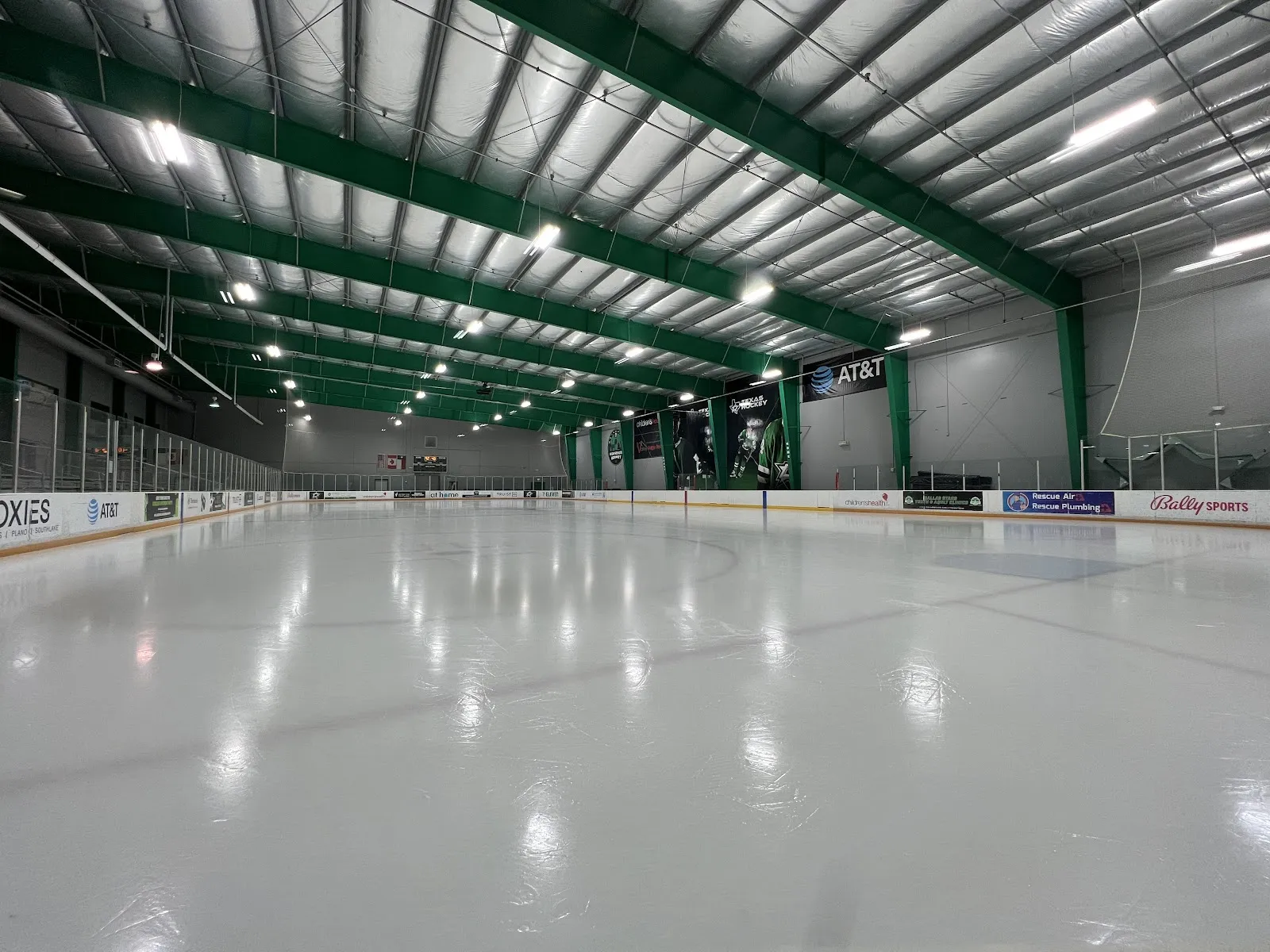 Empty ice sheet under green roof trusses, with AT&T, Bally Sports and other dasherboard ads, a 'Texas Hockey' banner and Dallas Stars player graphics on the end wall, and a 'childrenshealth' board. — Children's Health StarCenter Euless