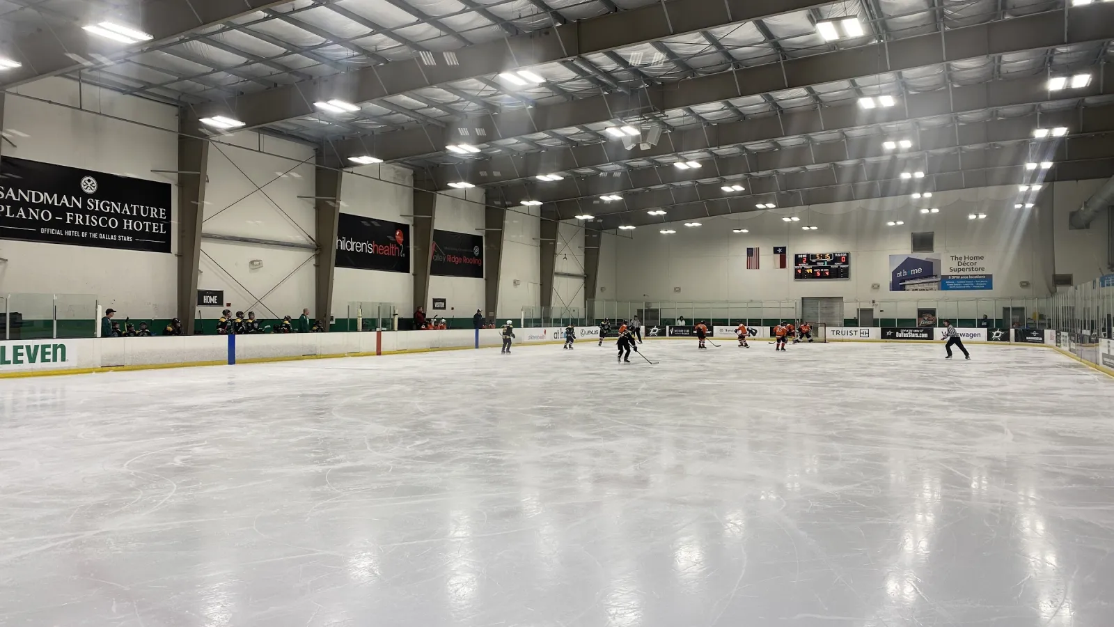 Un partido de hockey en el extremo lejano de una pista, visto desde las vallas cercanas, con mantas de patrocinadores en las paredes bajo un techo de acero verde. — Children's Health StarCenter Farmers Branch