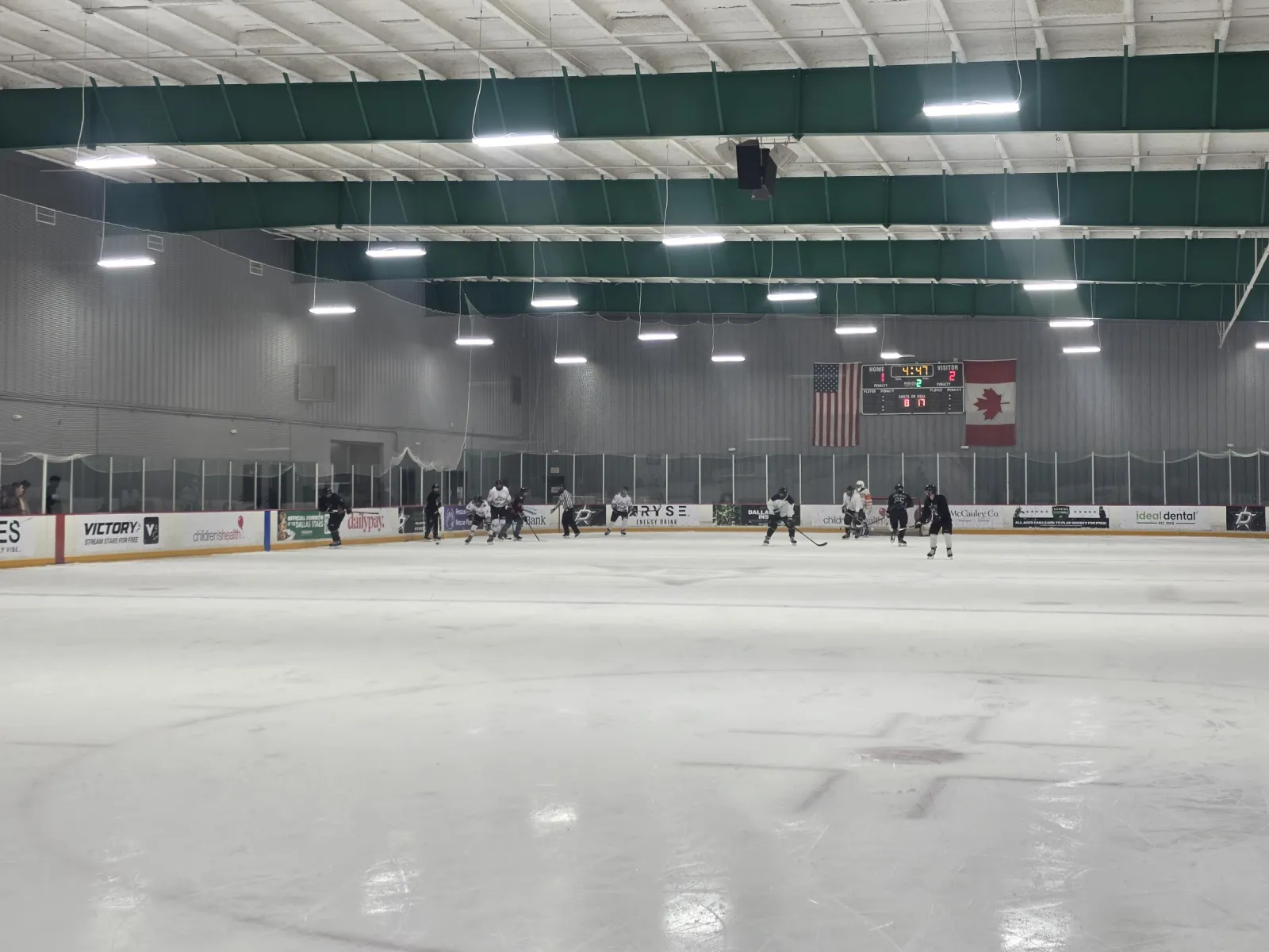 Todo el hielo visto desde un extremo, con jugadores en el extremo opuesto, el marcador entre banderas de Estados Unidos y Canadá y vigas verdes en el techo. — Children's Health StarCenter Richardson