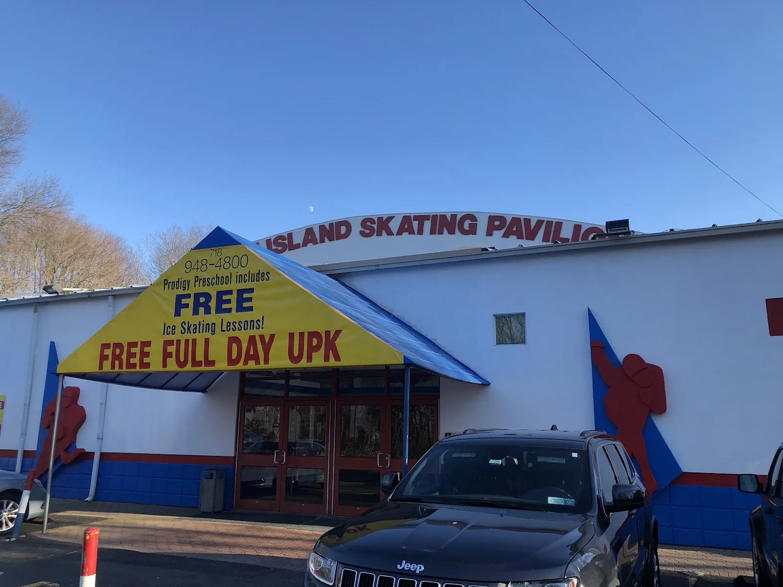 La entrada de Staten Island Skating Pavilion, un edificio blanco con figuras rojas de patinadores y su nombre a lo largo del borde del techo sobre un toldo — Staten Island Skating Pavilion