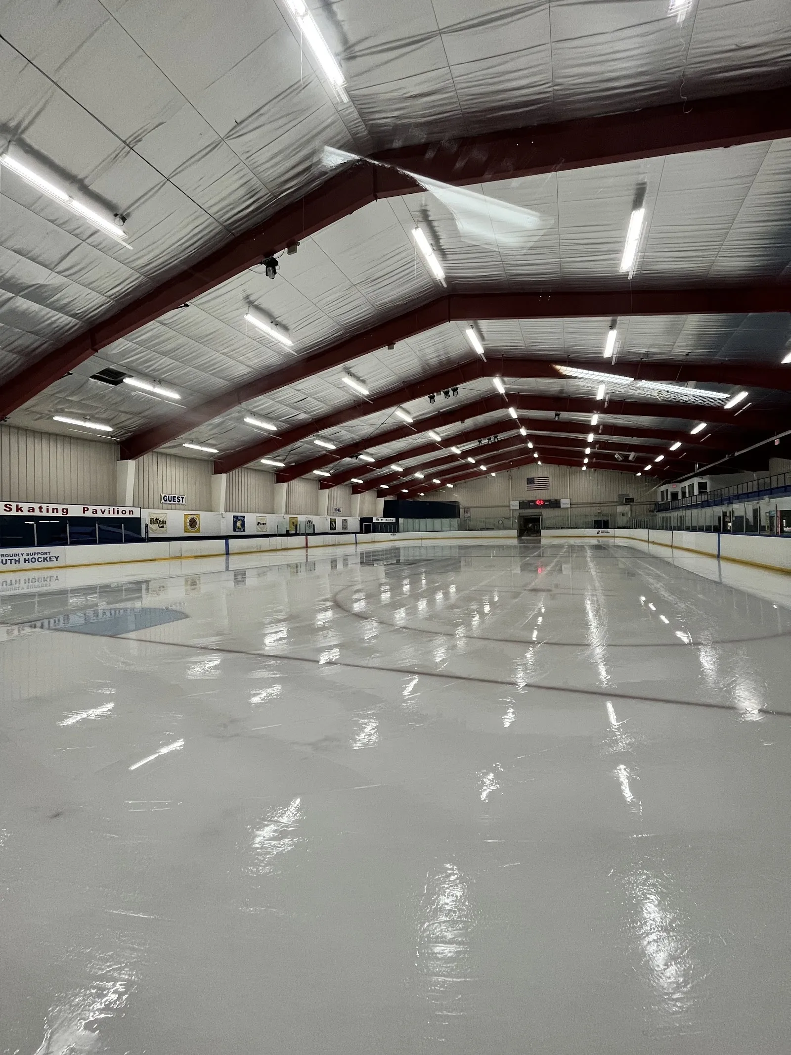 Una pista vacía bajo un largo techo con vigas de acero rojo oscuro, con el hielo blanco reflejando las luces a todo lo largo del recinto. — Staten Island Skating Pavilion