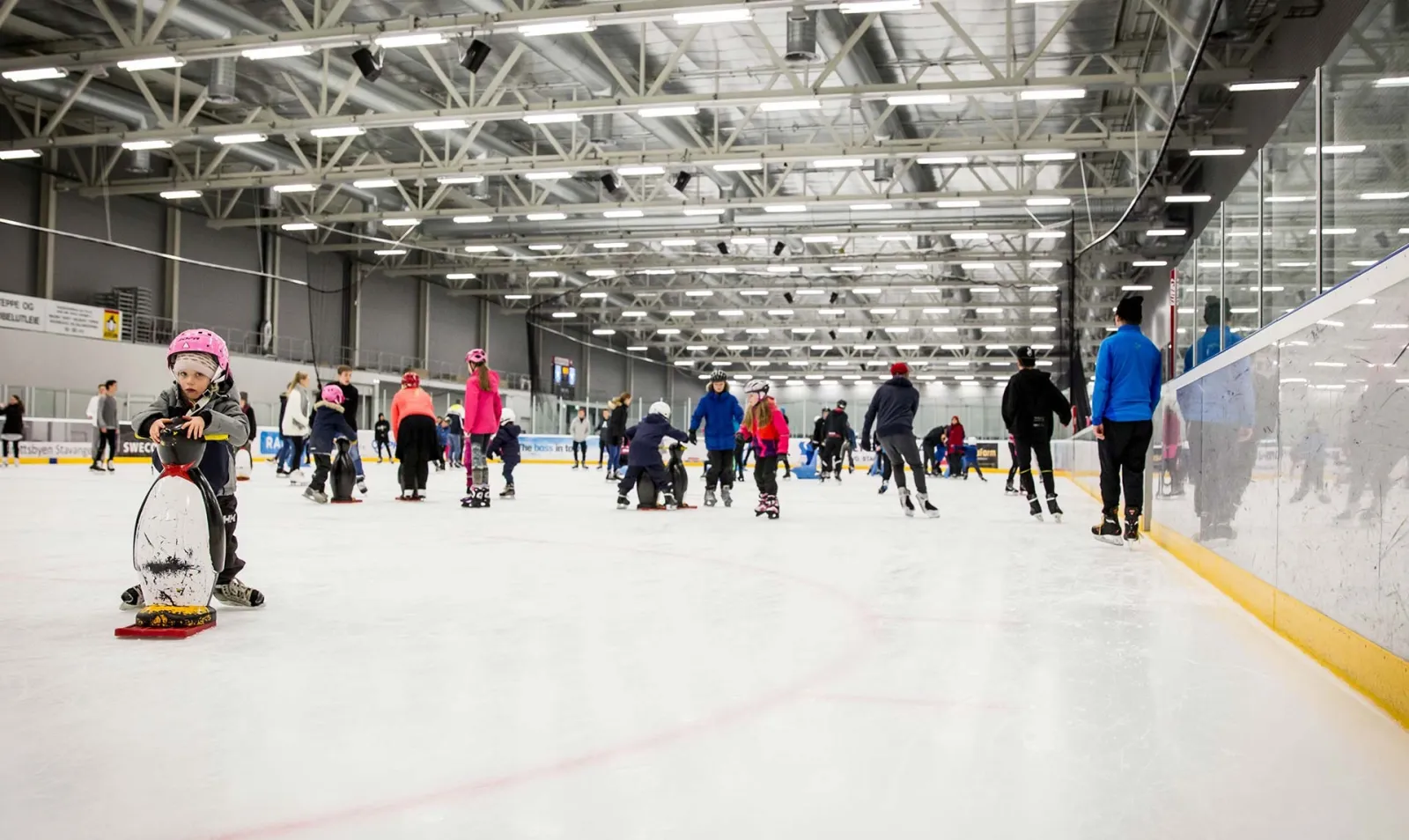 A public session in the Stavanger ice hall, skaters and children with penguin aids spread across the ice under the white steel roof. — Stavanger Ishall (Siddishallen)