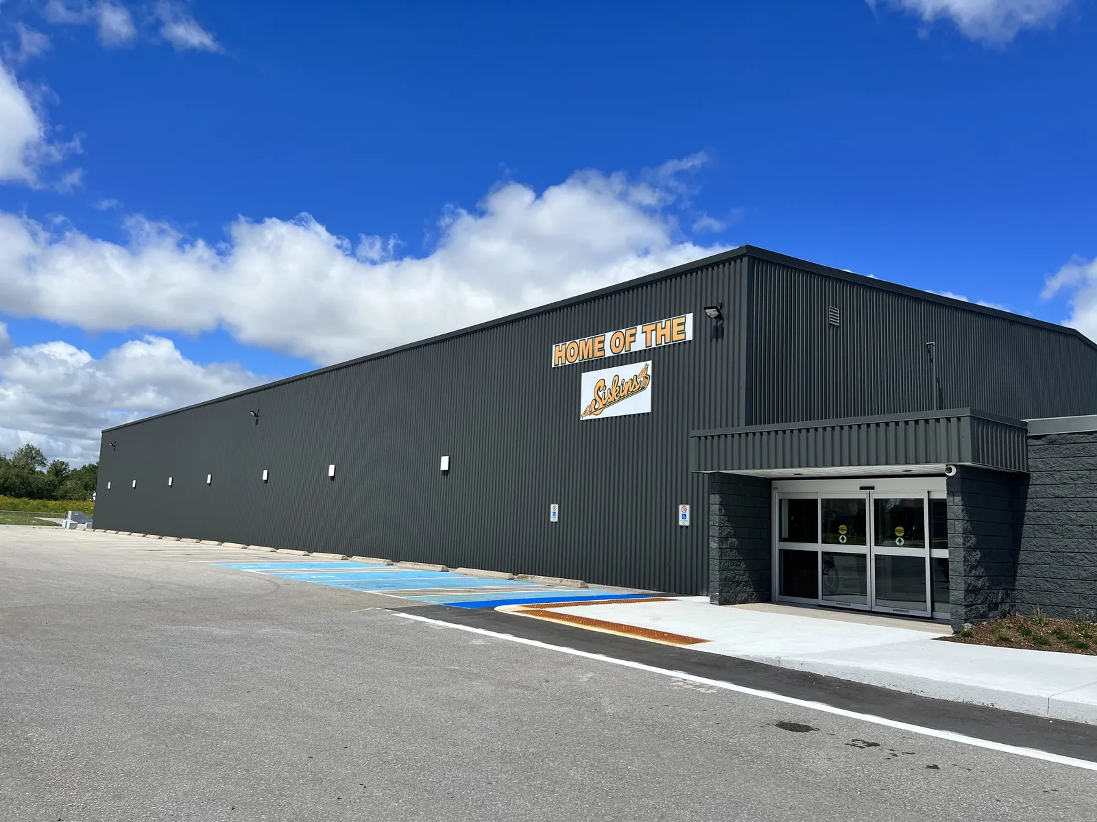 La entrada y el lateral del Stayner Arena, un edificio revestido de metal oscuro con las puertas correderas bajo un alero. — Stayner Arena