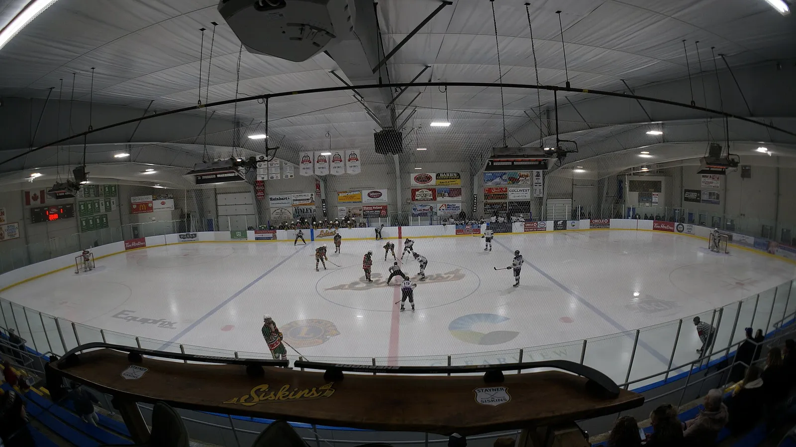 El hielo del Stayner Arena durante un partido de hockey, con espectadores junto a las bandas y carteles de patrocinadores en la pared del fondo. — Stayner Arena
