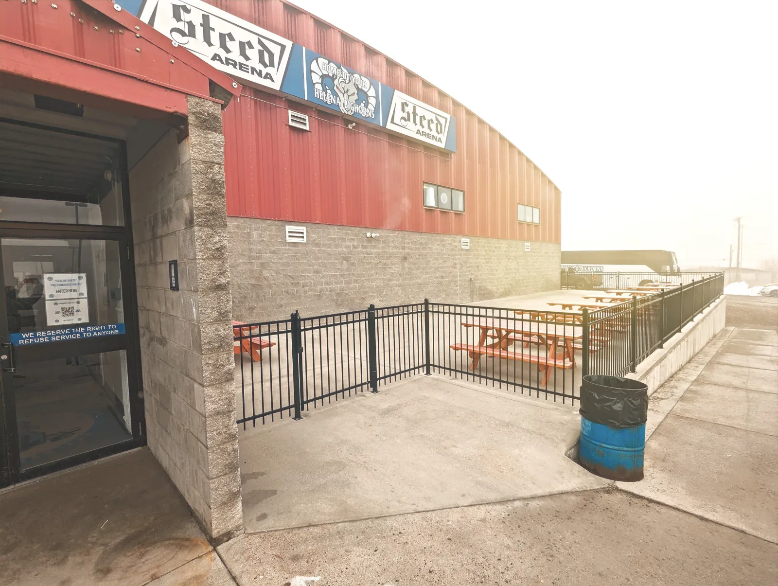 The side entrance of Steed Arena in Helena, with the red steel wall, the Steed lettering and the fenced patio with picnic tables — Steed Arena