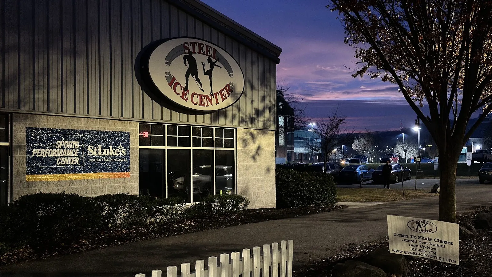 The Steel Ice Center at dusk, its oval skater sign on the grey metal wall above a lit window and hedges — The Steel Ice Center