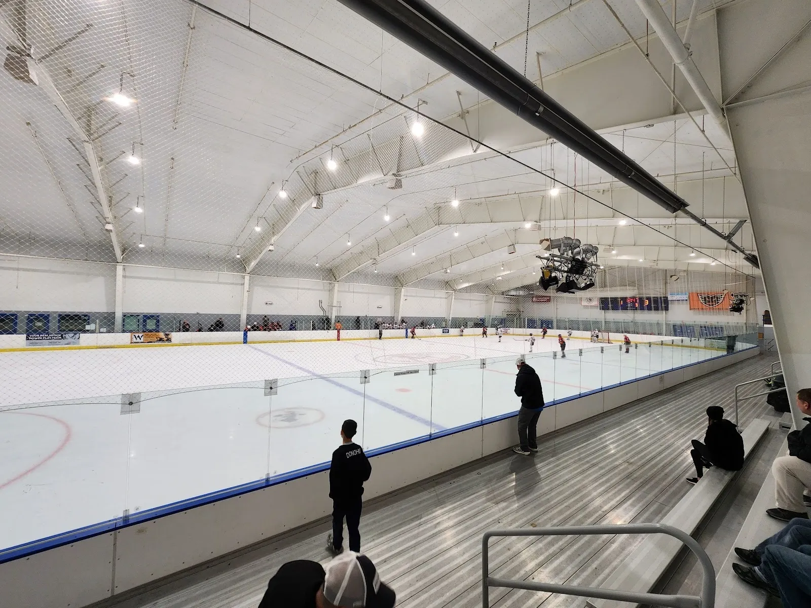 A wide sheet along a blue-lined rail in a white arched hall, a few people spread along the far boards and a faceoff circle on the ice. — The Steel Ice Center