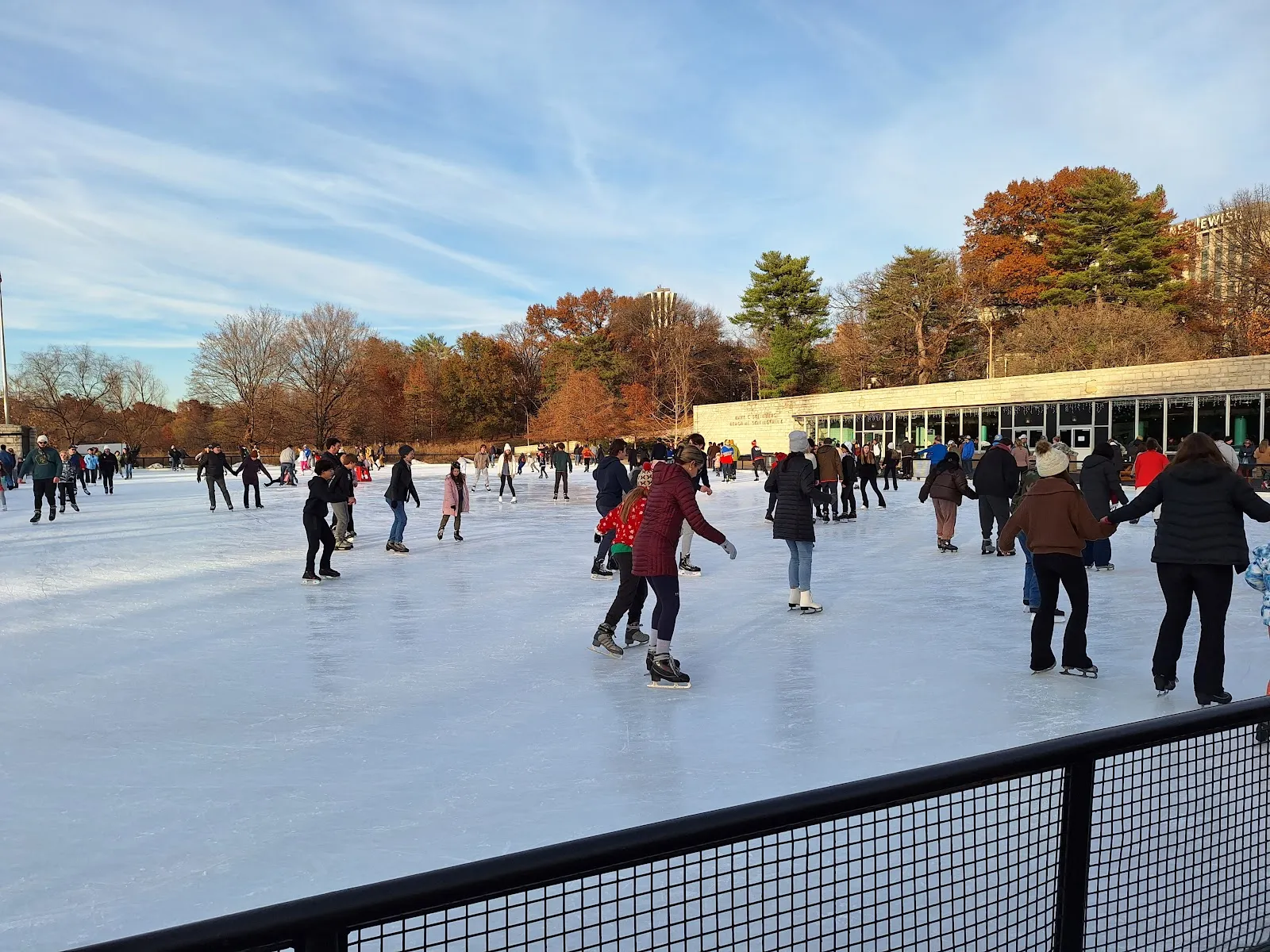Skaters circling the Steinberg rink on a winter afternoon, with fencing along the near edge and bare trees beyond. — Steinberg Rink