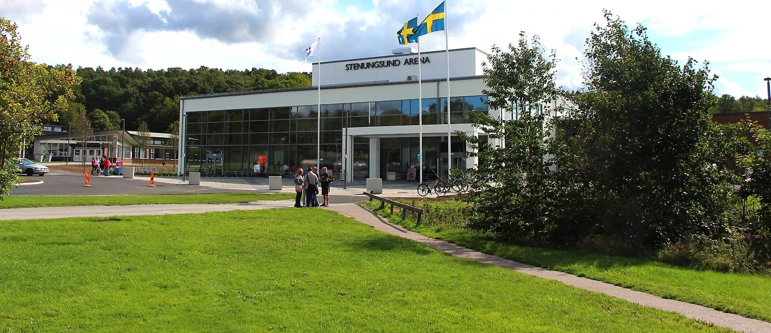 The glass-fronted Stenungsund Arena building with its name on the roof block, two Swedish flags, a lawn in front and a few visitors at the entrance. – Stenungsund Arena (ishall)