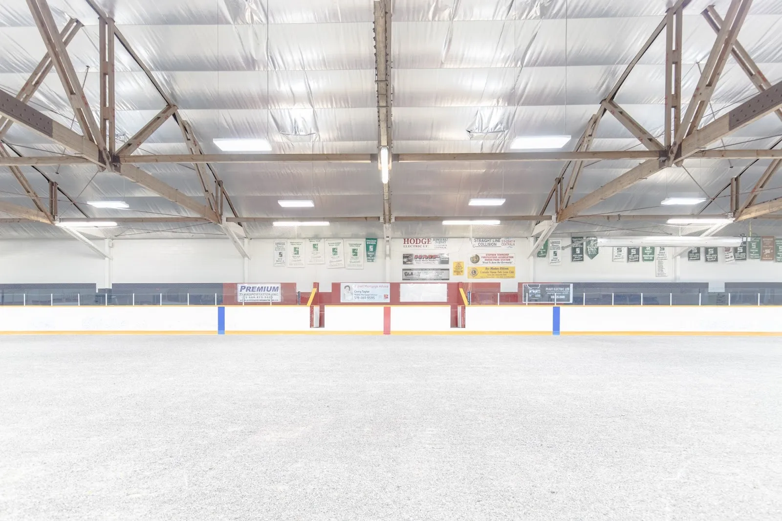 An empty sheet seen from the end, blue lines and red lines on white ice below a timber-trussed roof with light fittings. — Stephen Township Arena (Huron Park)