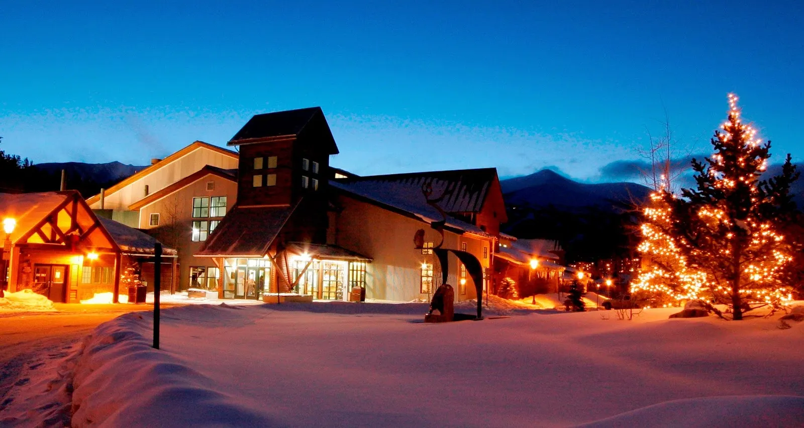 Dusk photo of Stephen C. West Ice Arena's front entrance, a timber-and-stucco building with a tall gabled tower, lit windows and doors, and a snow-covered plaza with a lit Christmas tree in front. — Stephen C. West Ice Arena