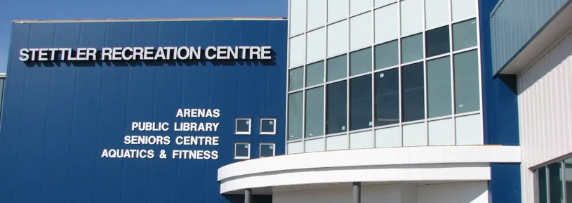 The blue facade of the Stettler Recreation Centre with its name and a sign listing arenas, public library, seniors centre and aquatics & fitness, beside a glass entrance. — Stettler Recreation Centre