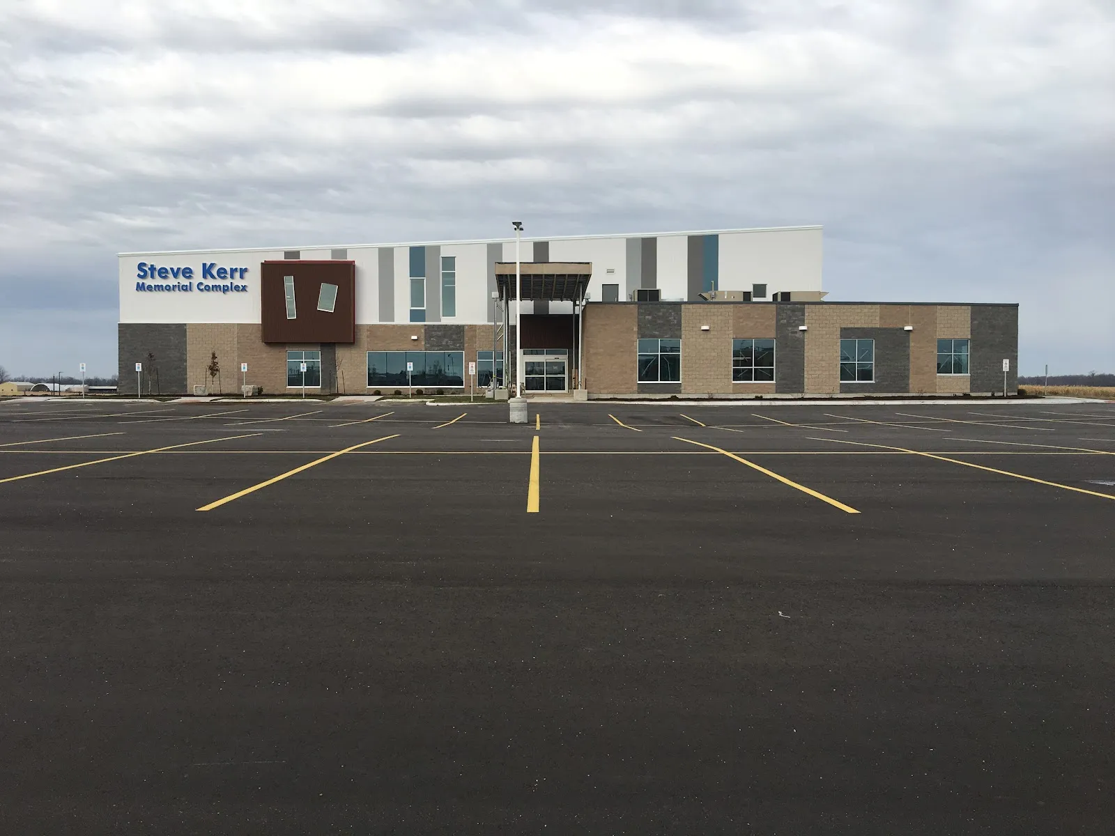 Steve Kerr Memorial Complex en Listowel, un edificio moderno de ladrillo, vidrio y paneles blancos con su nombre en el frente, visto desde el otro lado del estacionamiento. — Steve Kerr Memorial Complex (Listowel)