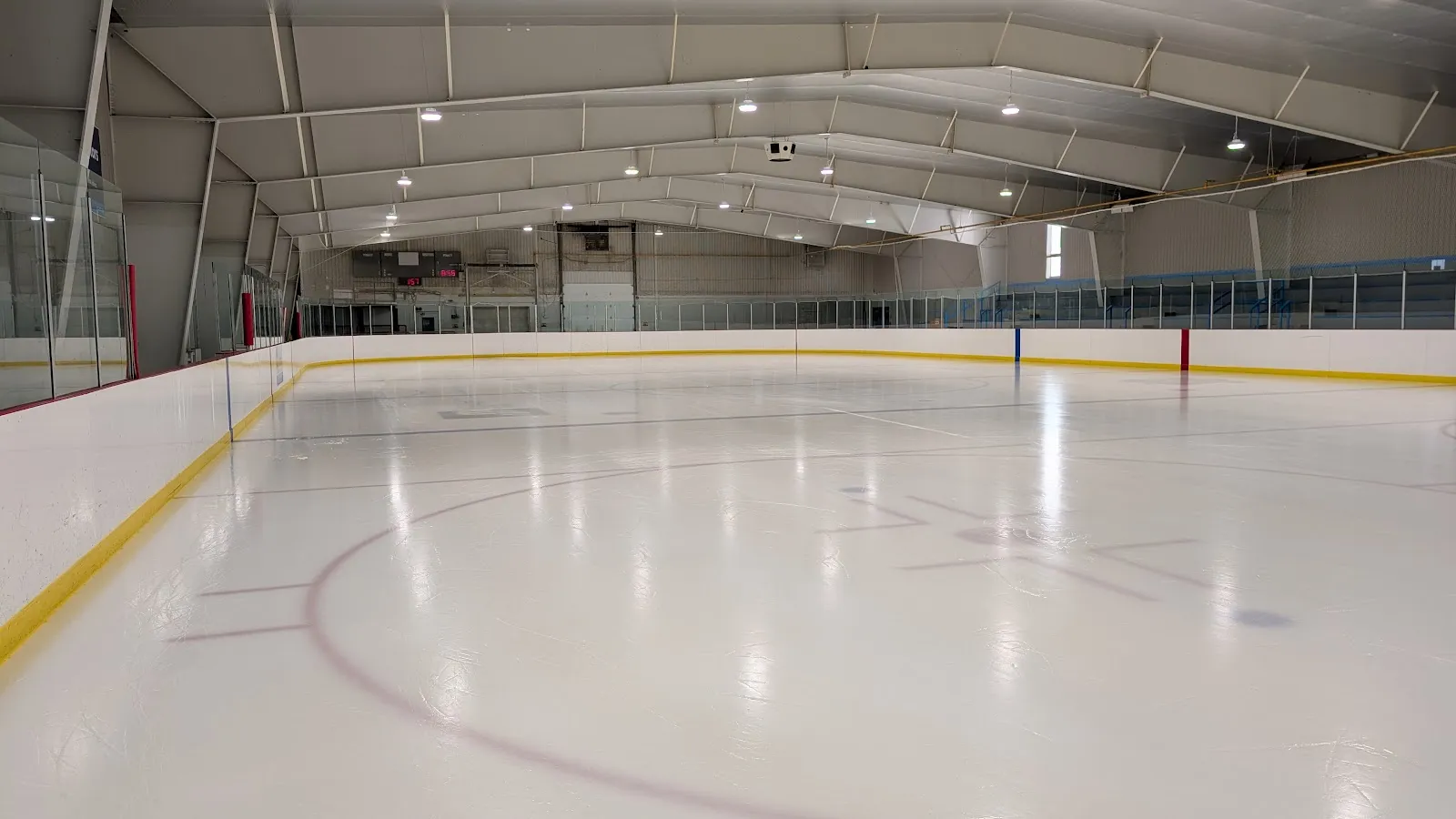 The empty ice from the corner, with yellow kickplates and a low white steel roof over the stands. — Stewart Community Centre (Pakenham)